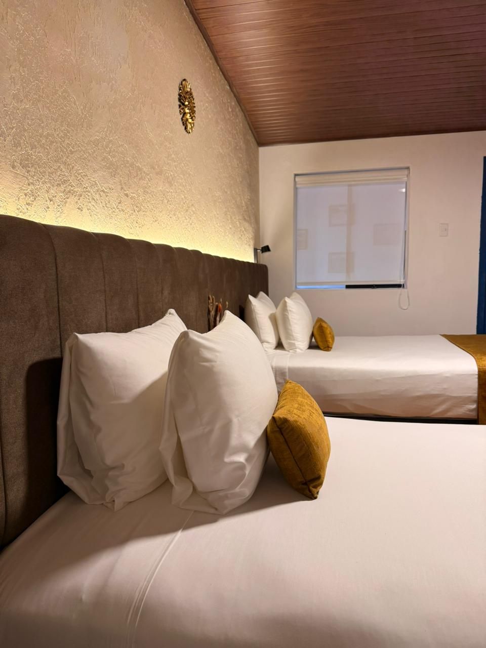 A hotel room with two beds, a wooden ceiling, a textured wall, and a window with blinds.