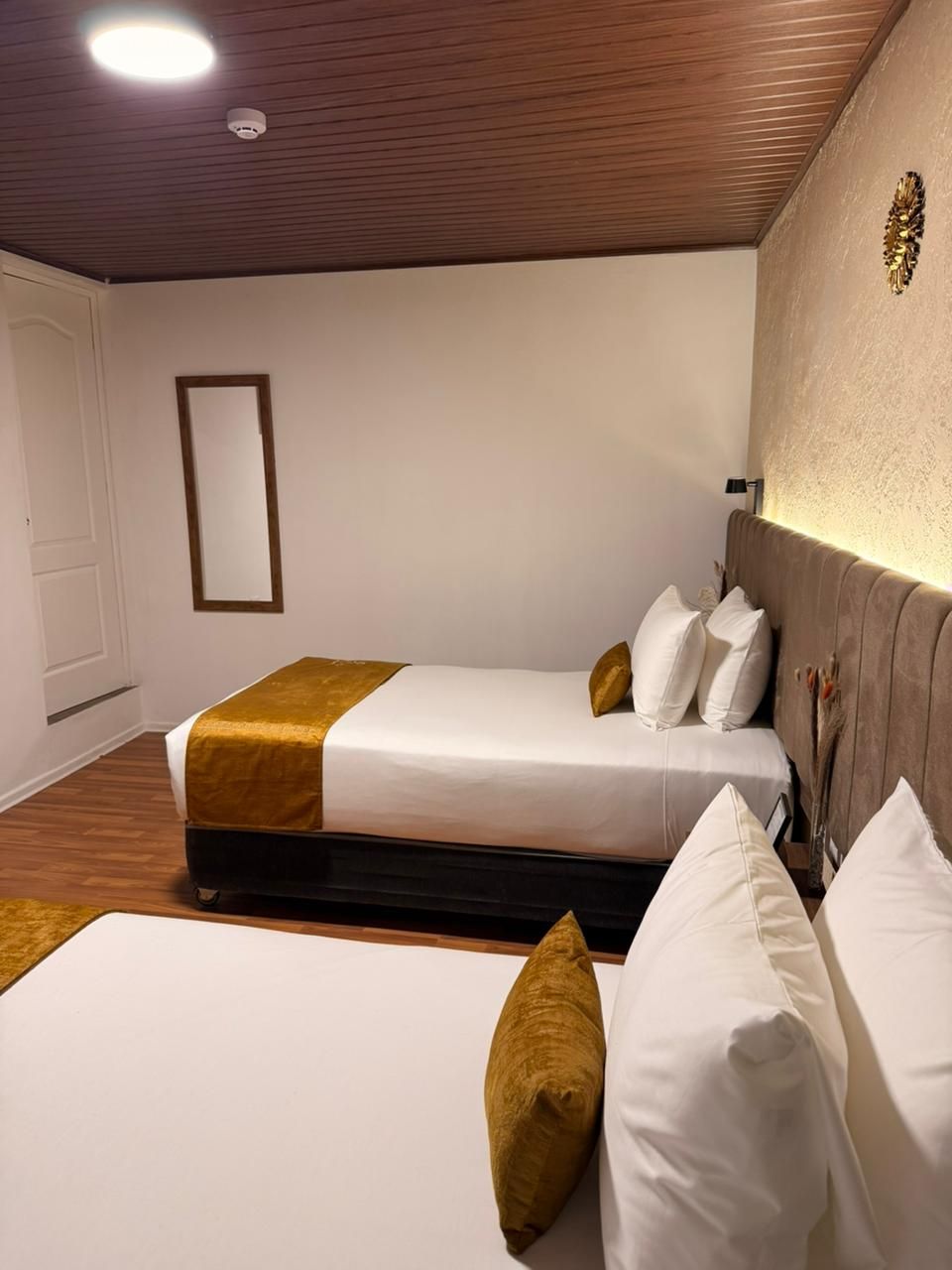A hotel room with two beds, wooden flooring, a mirror, and a wall-mounted light fixture.