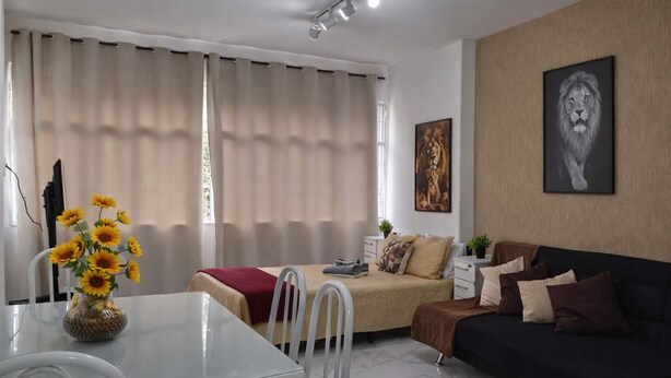 A modern living room with a dining area, featuring a glass table, white chairs, a dark sofa, and a vase of sunflowers. - Cozy Studio in the Heart of Rio (Near Lapa) (Rio de Janeiro)