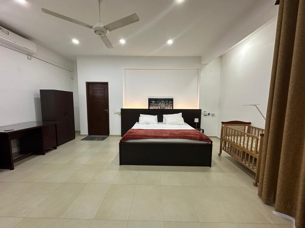 A bedroom with a bed, a ceiling fan, a TV, a desk, and a crib.