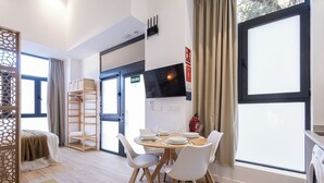 Ein modernes Hotelzimmer mit Essbereich, einem Bett mit Holz-Kopfteil, einem Kleiderschrank und einem großen Fenster mit Vorhängen.