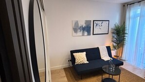A modern living room with a dark blue sofa, two framed abstract paintings, a round mirror, a small black coffee table with glasses, and a potted plant. - Garden Suite Paris Alfortville Metro - 10 min from Paris (Alfortville)