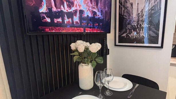 A dining area with a TV displaying a fire scene, a framed picture, and a vase with white flowers. - Garden Suite Paris Alfortville Metro - 10 min from Paris (Alfortville)