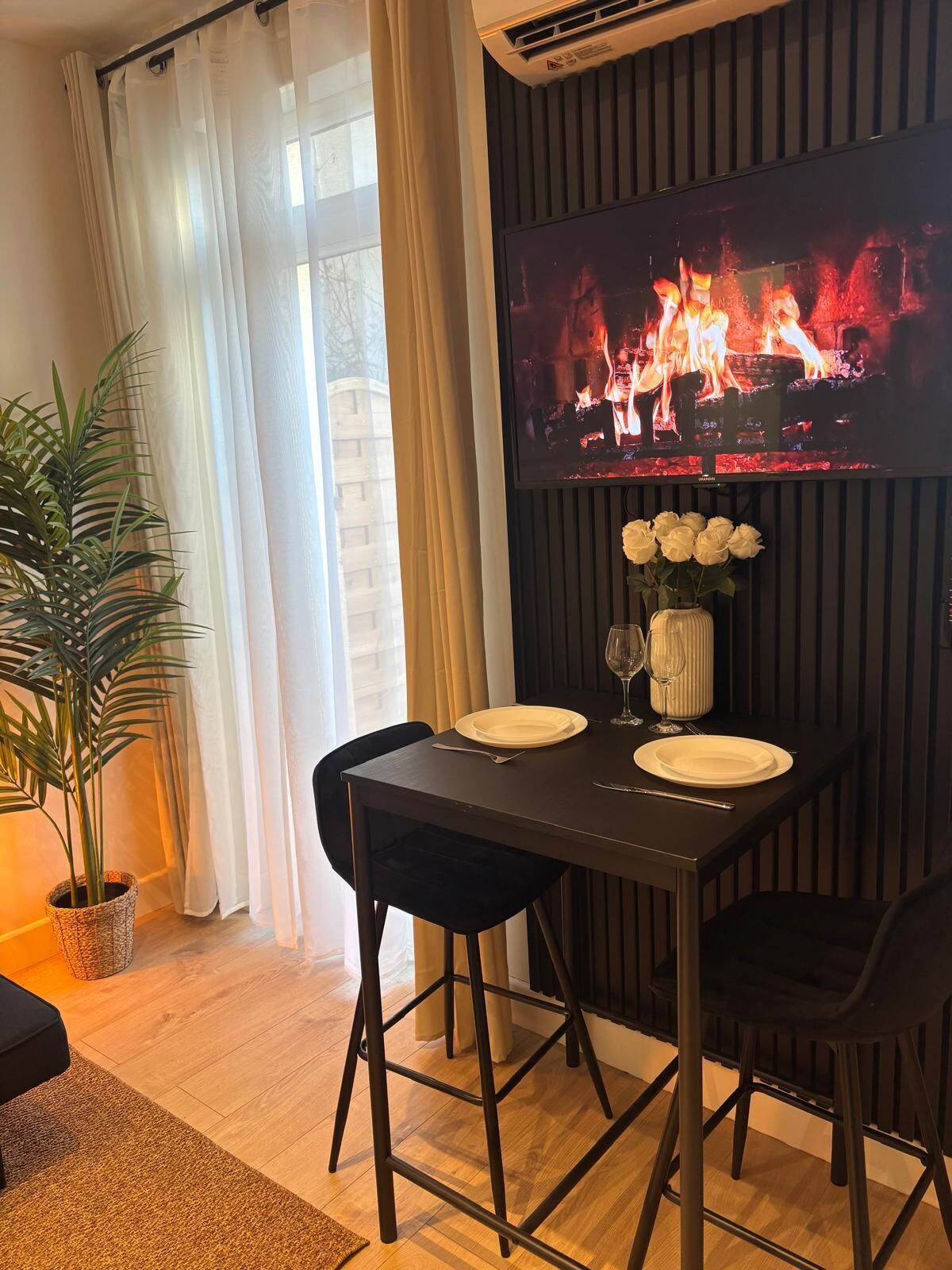 A dining area with a TV displaying a fireplace, a table set with plates and a vase of flowers, and a potted plant.