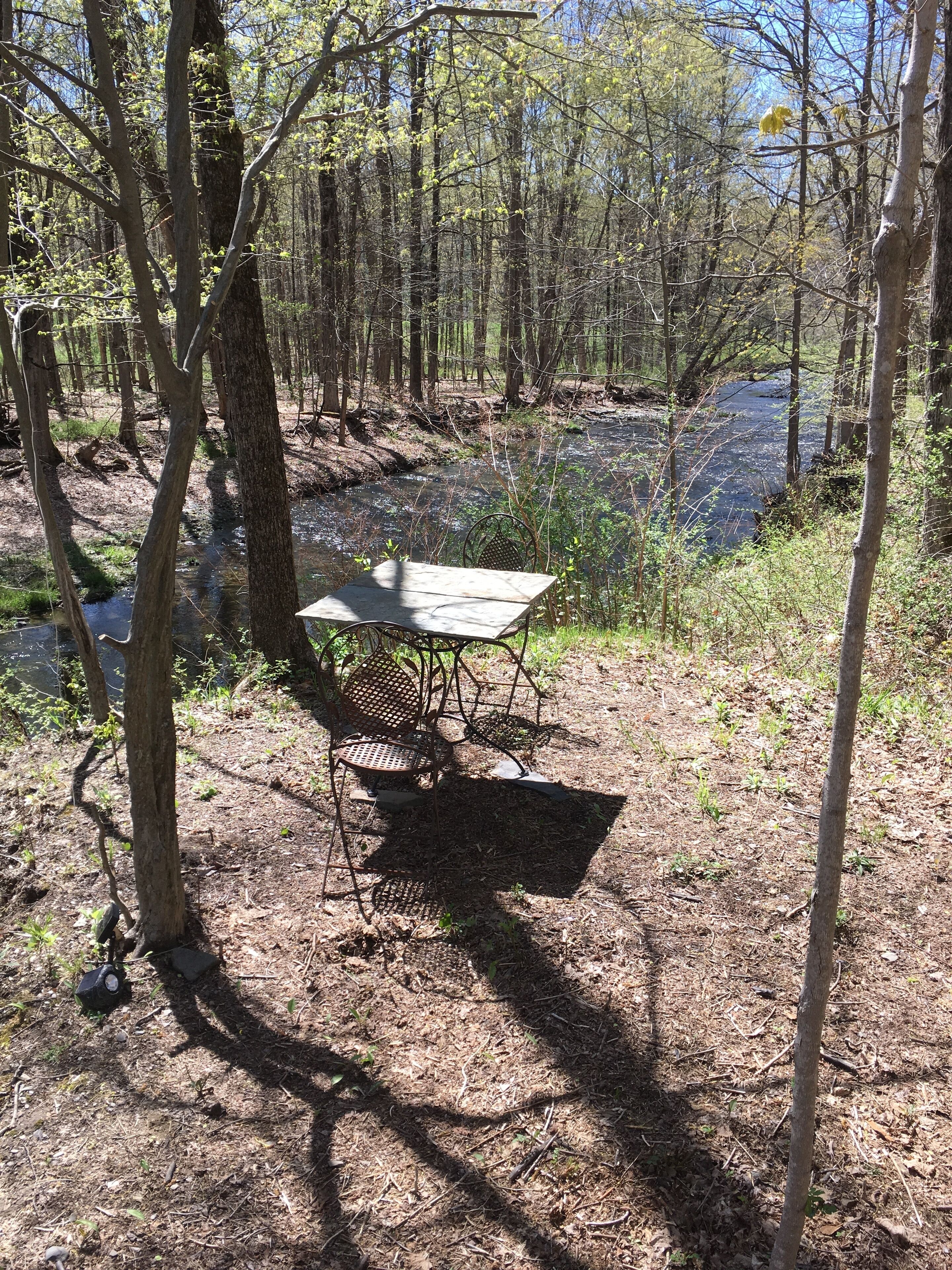 A picnic table with metal chairs in a forested area near a stream.