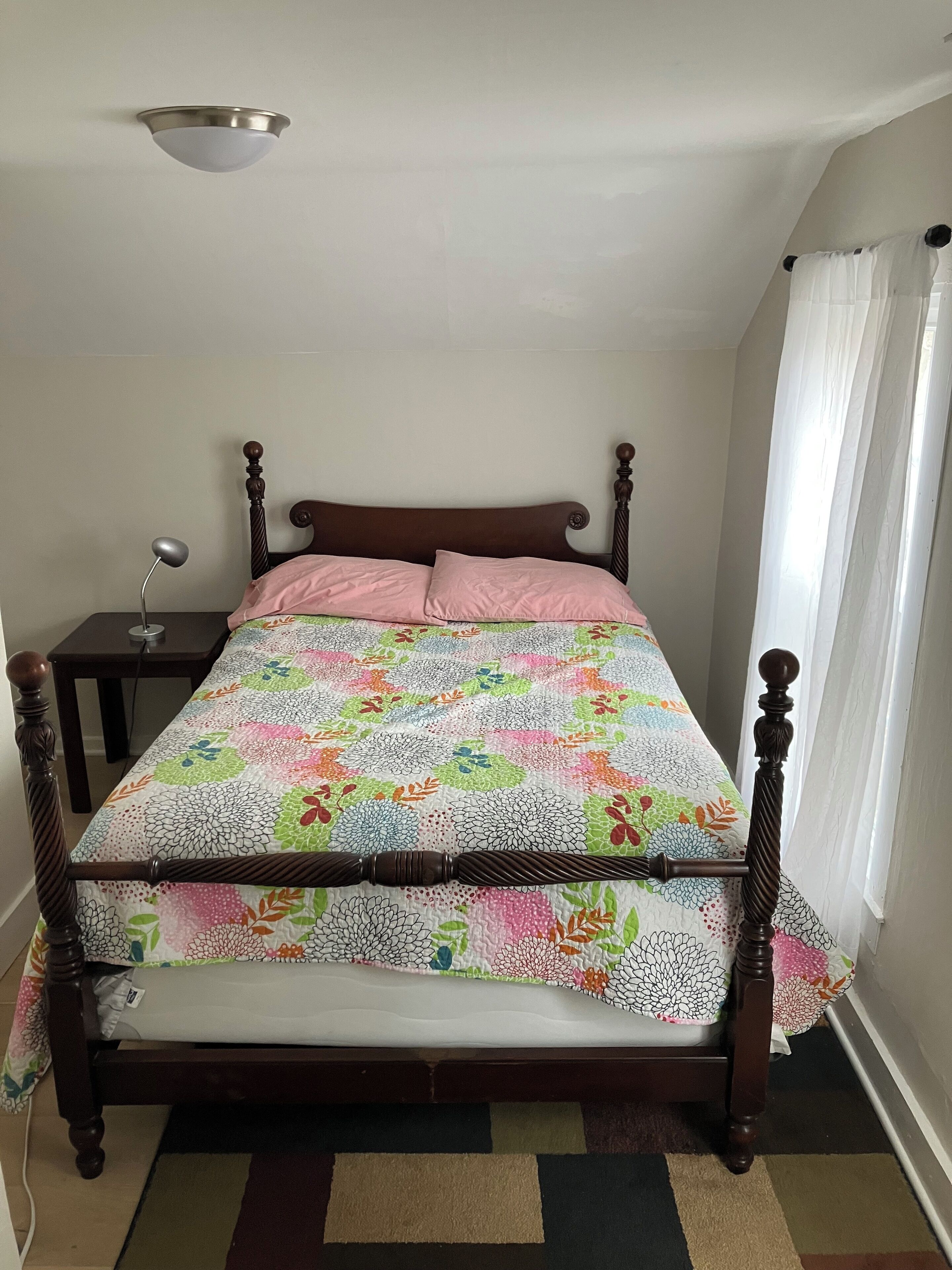 Un letto singolo con struttura in legno, una coperta colorata, un comodino con una lampada e un lampadario al soffitto.