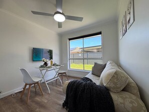 Een woonkamer met een bank, een eettafel met stoelen, een plafondventilator en een groot raam met uitzicht op een schutting en bomen.