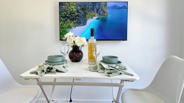 A dining table with a vase of white flowers, a bottle, and two glasses. A flat-screen TV displays a scenic coastal image. - Modern Belmont Suite Easy access to CBD & Bayside (Belmont)
