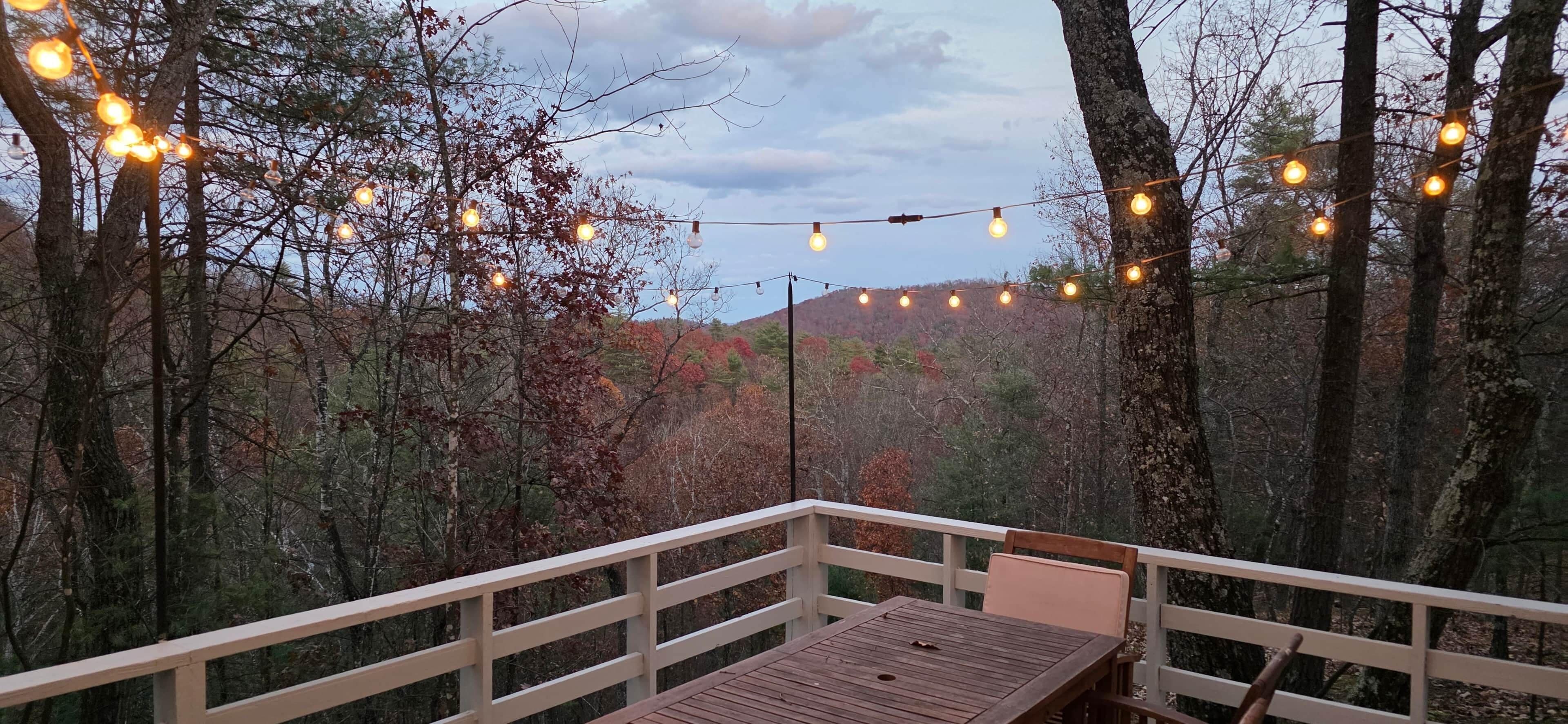 A wooden deck with a table and chairs, surrounded by trees and string lights.