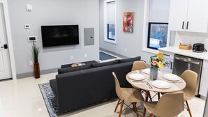 A modern living room with a black sofa, a flat-screen TV, and a dining area with a table set for two. - 20 Min to Times Square-Stylish 2 BR w/Parking (West New York)