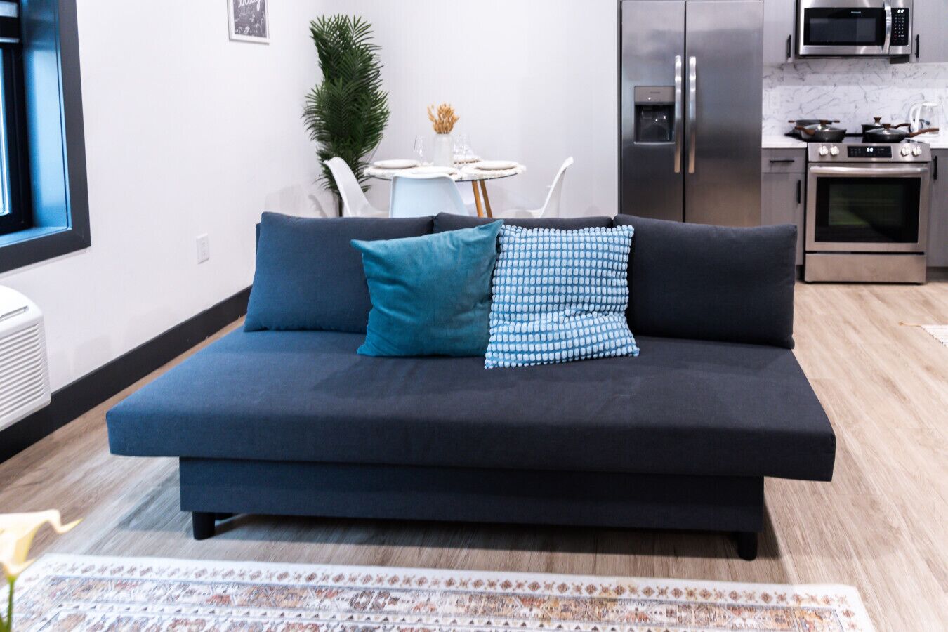 A modern living room with a dark sofa, a patterned throw pillow, a small dining table, and a kitchen area with appliances.