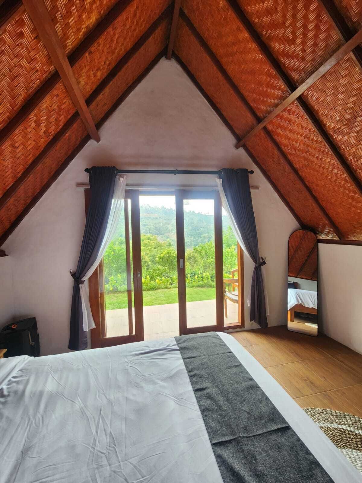 A bedroom with a large bed, a view of greenery through a sliding door, and a sloped ceiling with wooden beams.