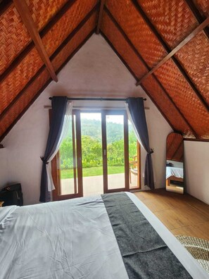 A bedroom with a large bed, a view of greenery through a sliding door, and a sloped ceiling with wooden beams. - Pondok Kanginan (Pelaga)