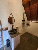Un baño con lavabo de piedra, un lavamanos de madera, un espejo montado en la pared, una ducha con fondo de panel de madera y un estante pequeño con artículos de aseo.