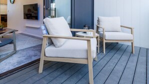 To lyse stole med hynder på en træterrasse, et lille rundt bord med kopper og en elkedel, samt et fjernsyn ophængt på væggen.