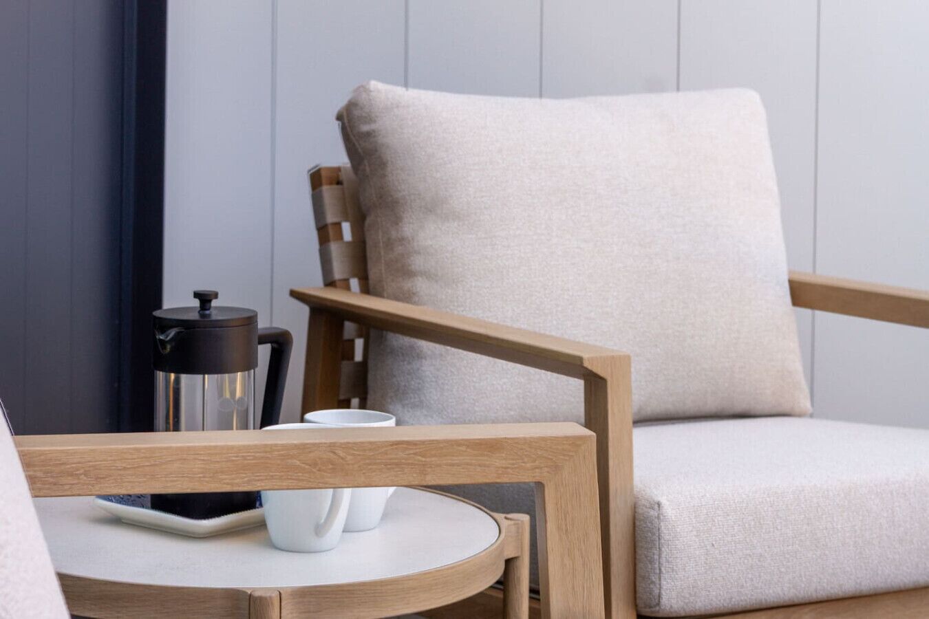 Una silla de madera con cojín, una mesita redonda pequeña con una taza blanca y un termo, y una cafetera negra.