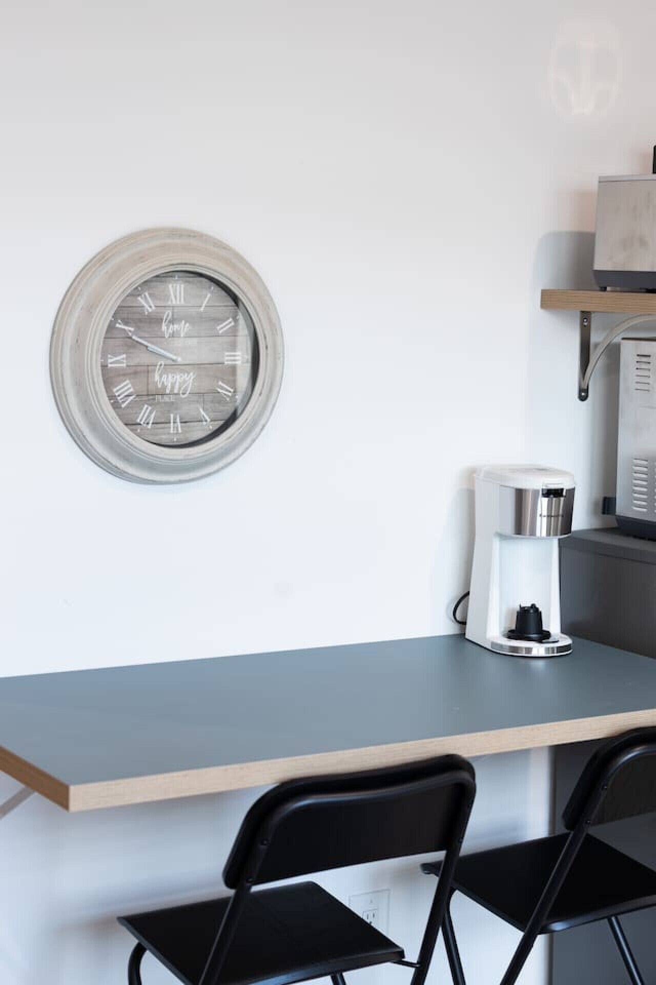 A kitchen area with a round wall clock, a coffee maker, and two black chairs.