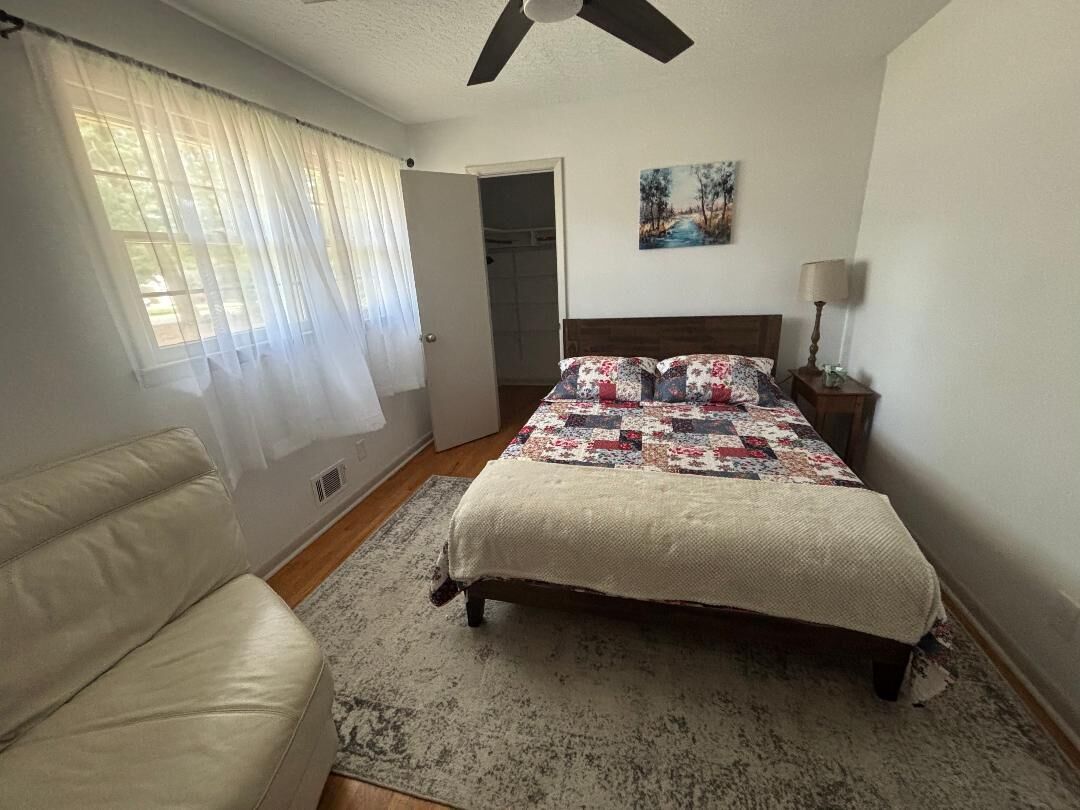 A bedroom with a bed, a chair, a ceiling fan, a painting, and a door.