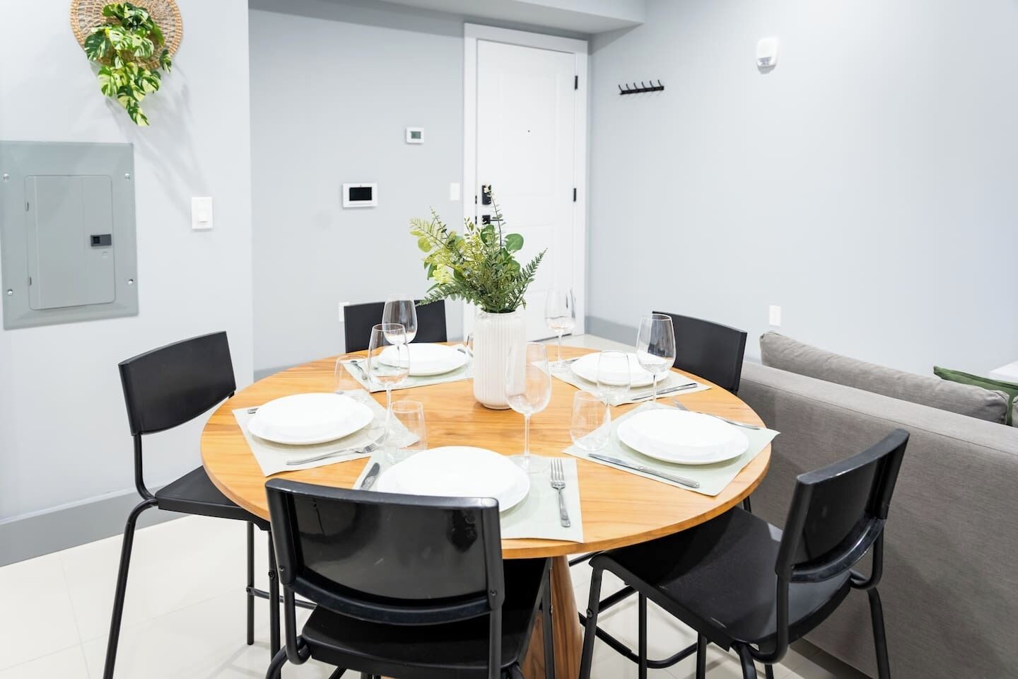 A dining area with a round wooden table set for four, surrounded by black chairs. A potted plant is on the table, and there is a whiteboard on the wall.