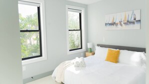 Un dormitorio con una cama grande, dos ventanas, un cuadro y una mesita de noche.
