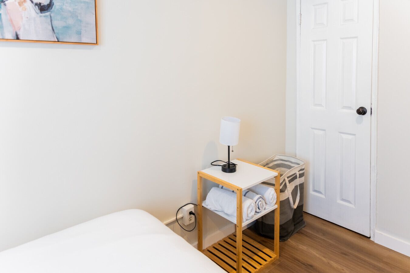 A hotel room with a bed, a bedside table with a lamp, a suitcase, and a trash bin.