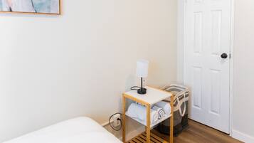 A hotel room with a bed, a bedside table with a lamp, a suitcase, and a trash bin.