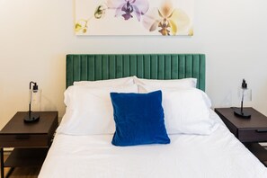 Un lit bien fait, avec du linge de lit blanc et un oreiller bleu, flanqué de deux tables de chevet avec des lampes, dans une chambre dont la tête de lit est recouverte d’un tissu vert.