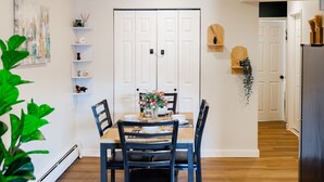 A dining area with a white double door, a table set with plates, cups, and a vase of flowers. There are shelves with decorative items and a plant in the foreground.