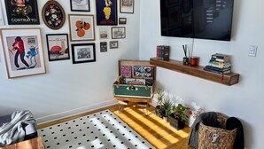 A living room with a wooden floor, a patterned rug, a television mounted on the wall, and various framed artworks and books on shelves.