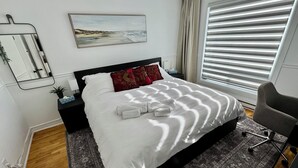 A neatly made bed with white linens and towels, a bedside table with a lamp, a nightstand, a painting on the wall, and a window with blinds.