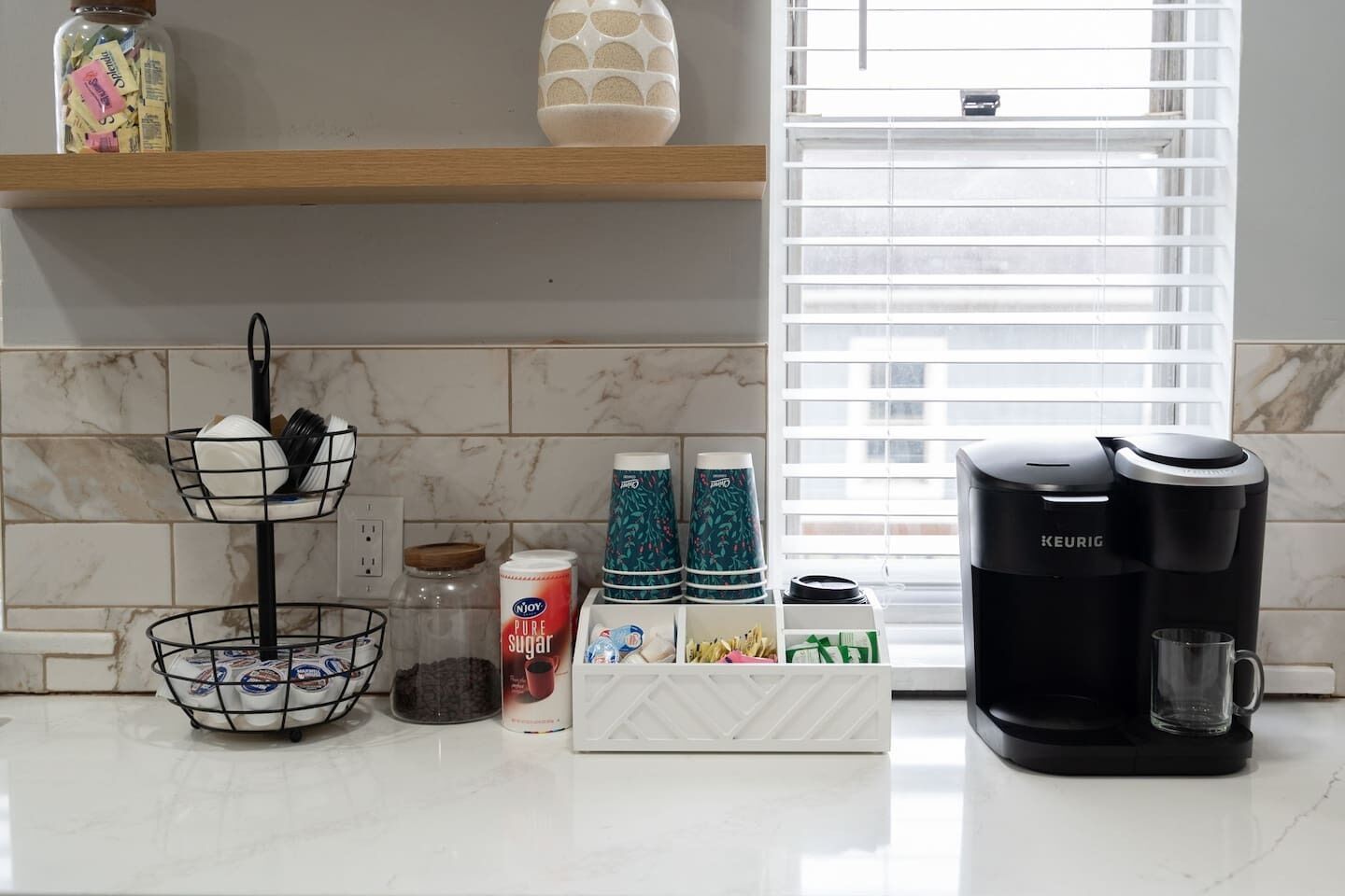 Et køkken med marmor bordplade, en Keurig kaffemaskine, en hvid kop, et krydderihold og et sukkerglas.