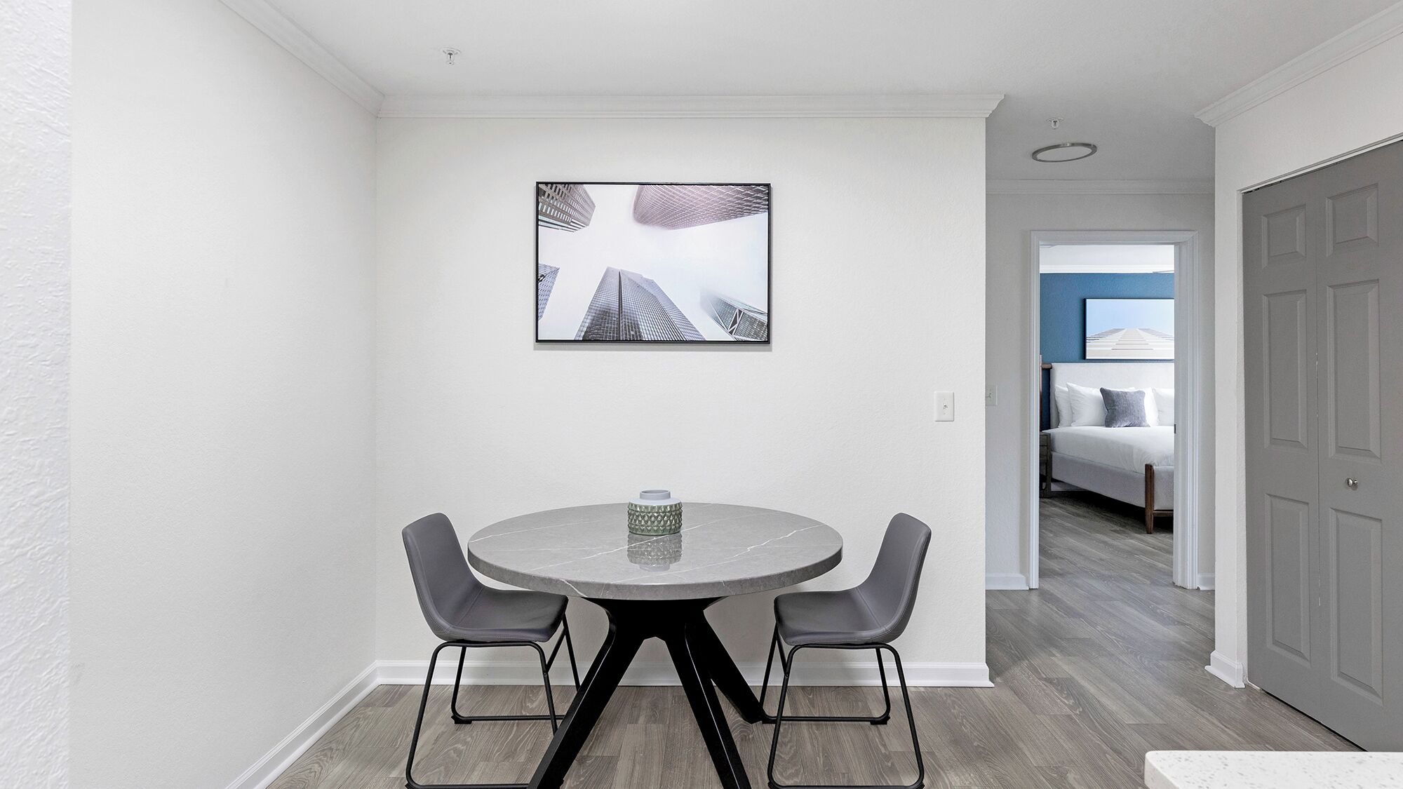Uma sala de jantar moderna com uma mesa redonda e duas cadeiras, uma obra de arte emoldurada na parede, e vista para um quarto com uma cama e uma mesa de cabeceira.