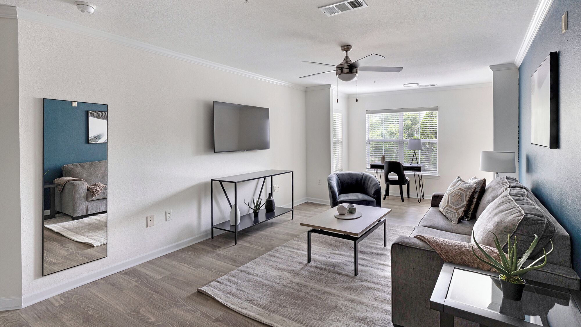 A modern living room with a flat-screen TV, a ceiling fan, a sofa, a coffee table, and a large window with blinds.