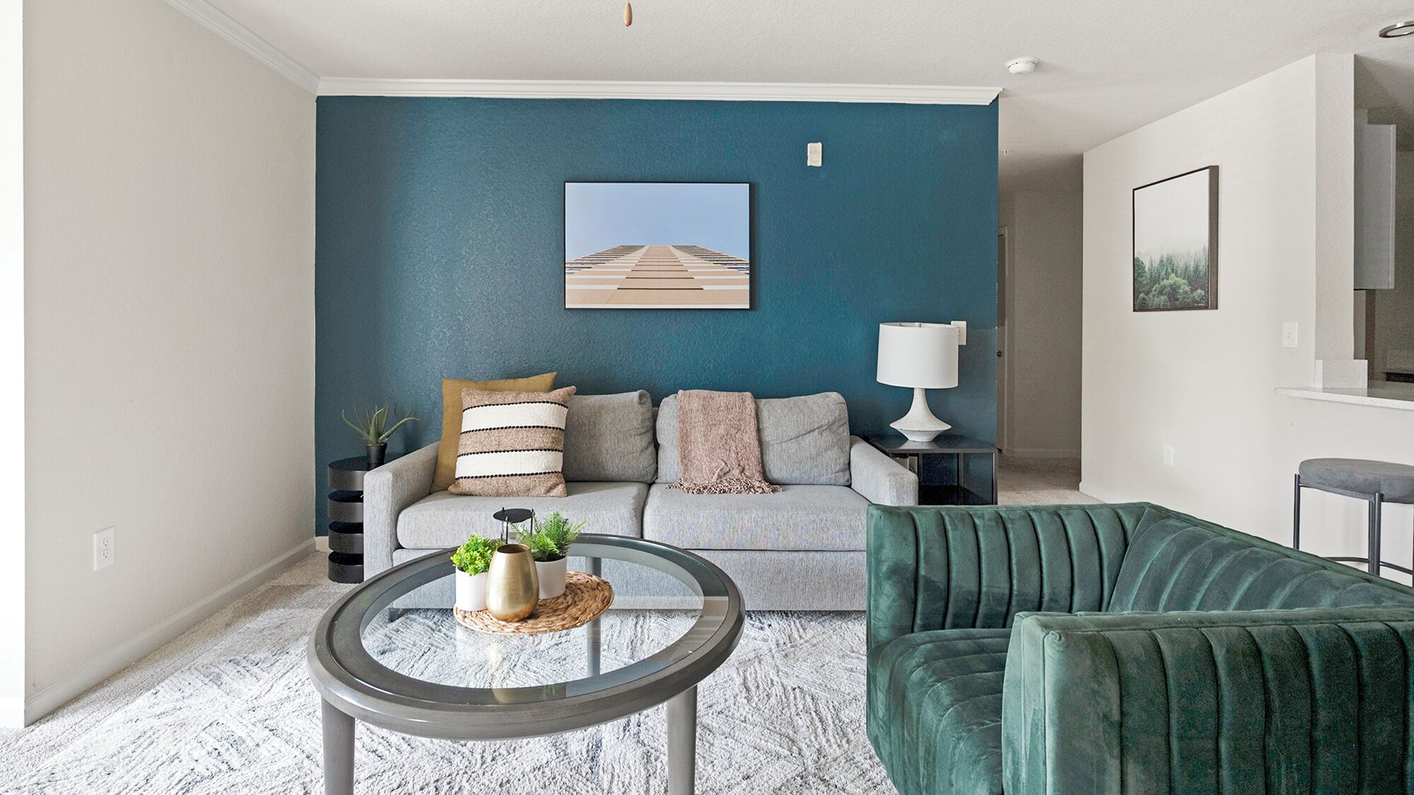 A living room with a teal accent wall, a sofa with striped pillows, a green velvet armchair, a glass coffee table with decorative items, and a framed picture above the sofa.