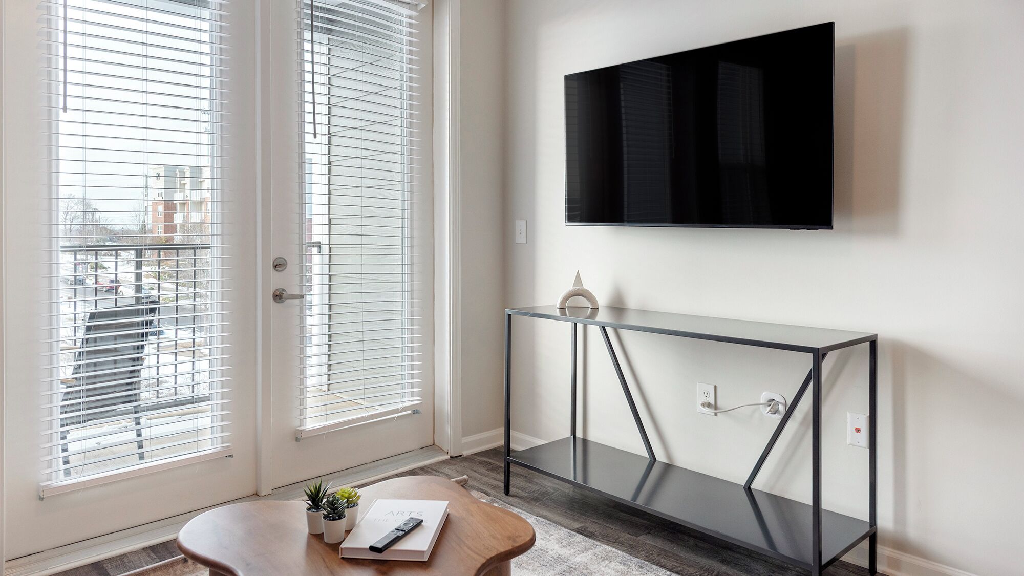 Ein modernes Zimmer mit einem Glastischchen, einem an der Wand montierten Fernseher und einer Tür mit Jalousien.