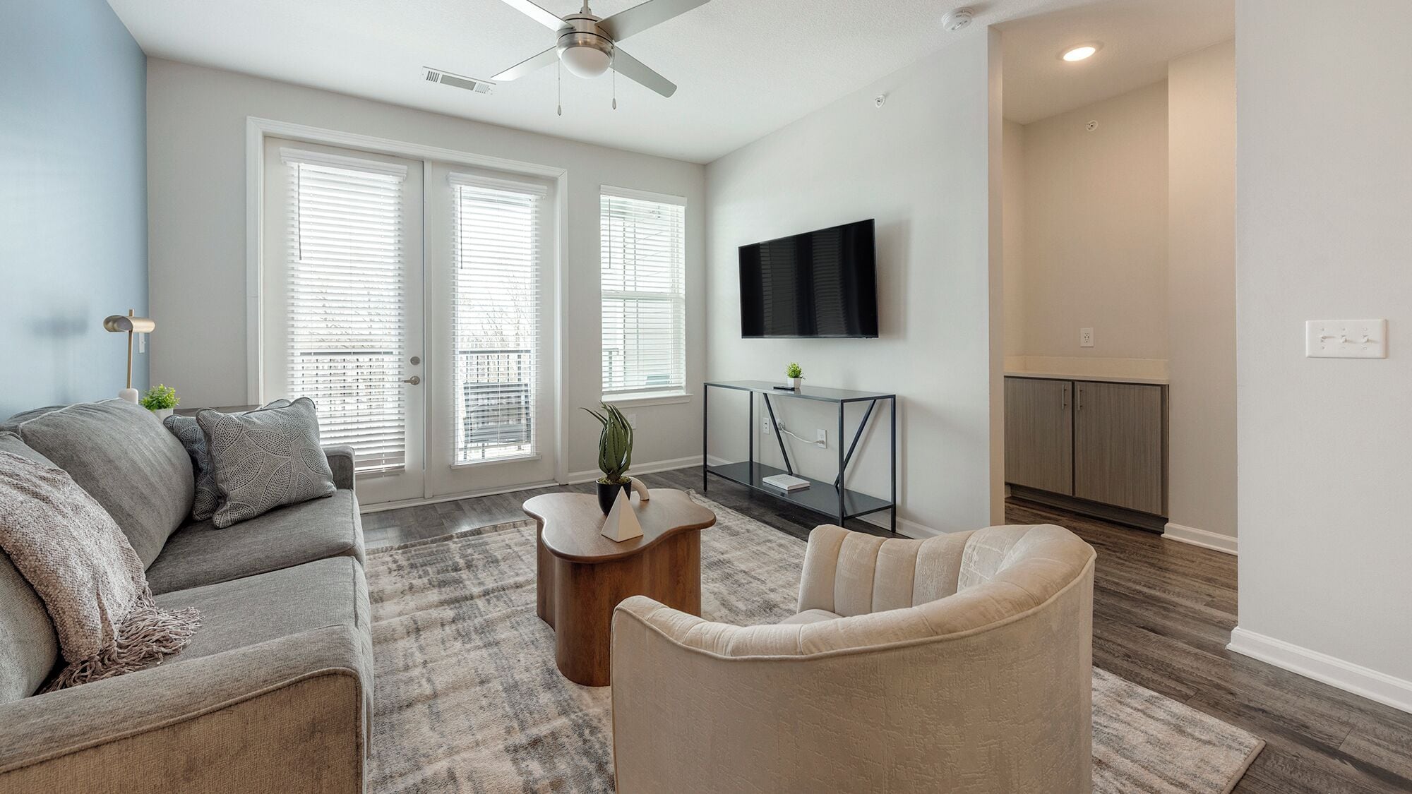 Una sala de estar moderna con un sofá gris, un sillón beige, una mesa de centro y un televisor montado en la pared.