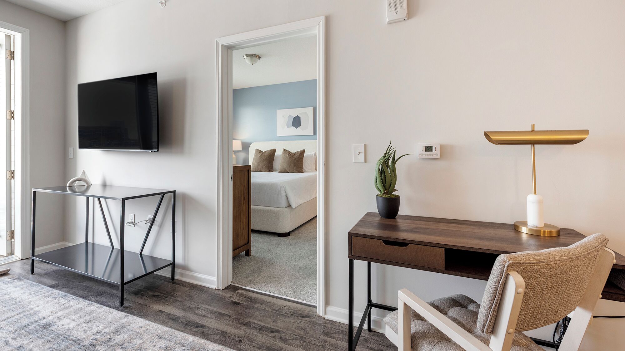 A modern hotel room with a flat-screen TV, a desk with a lamp, a chair, and a potted plant.