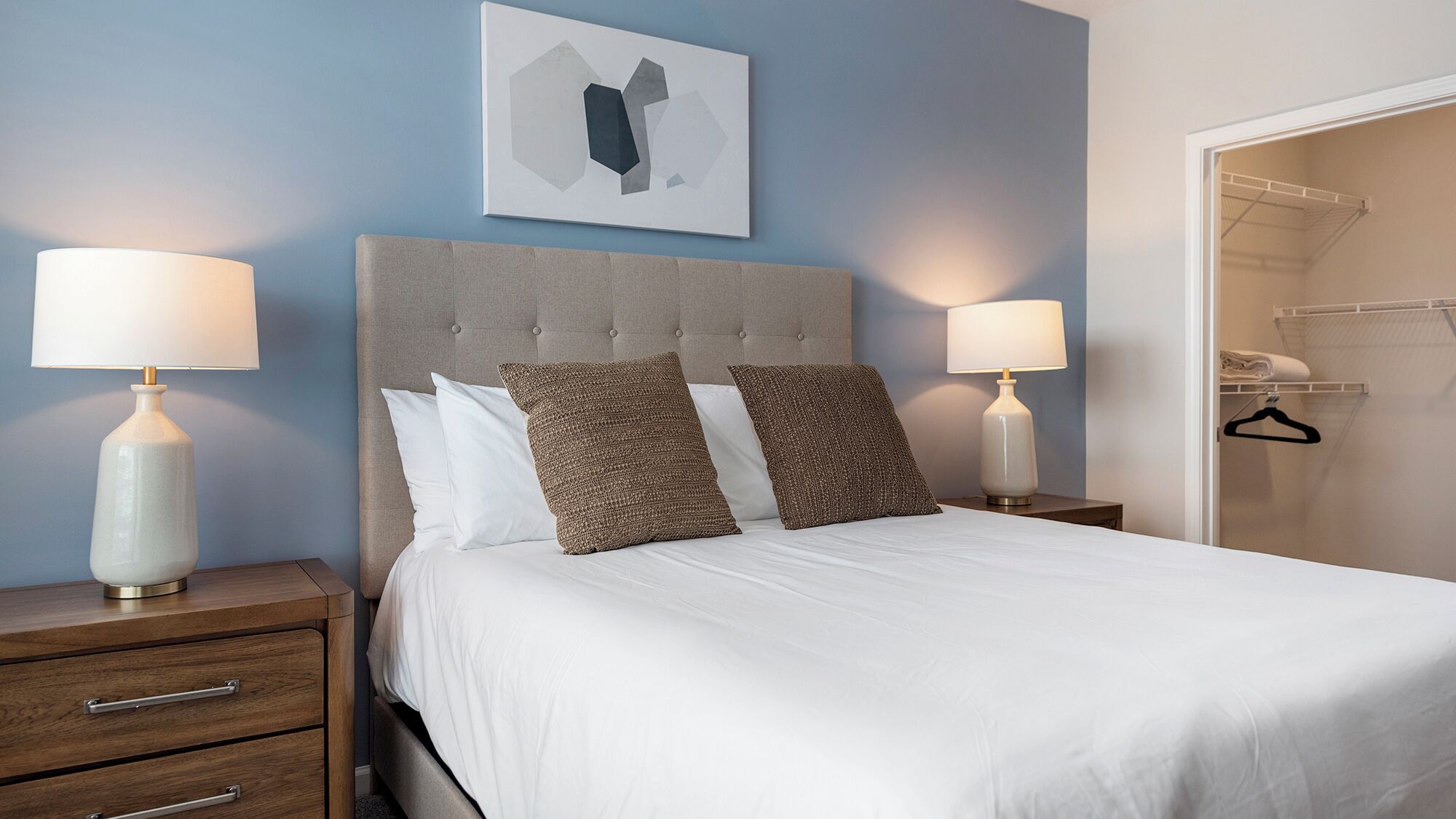 Cama bem arrumada com lençóis brancos e duas almofadas texturizadas, com mesas de cabeceira com luminárias de cada lado, em frente a uma parede azul com uma obra de arte geométrica acima.