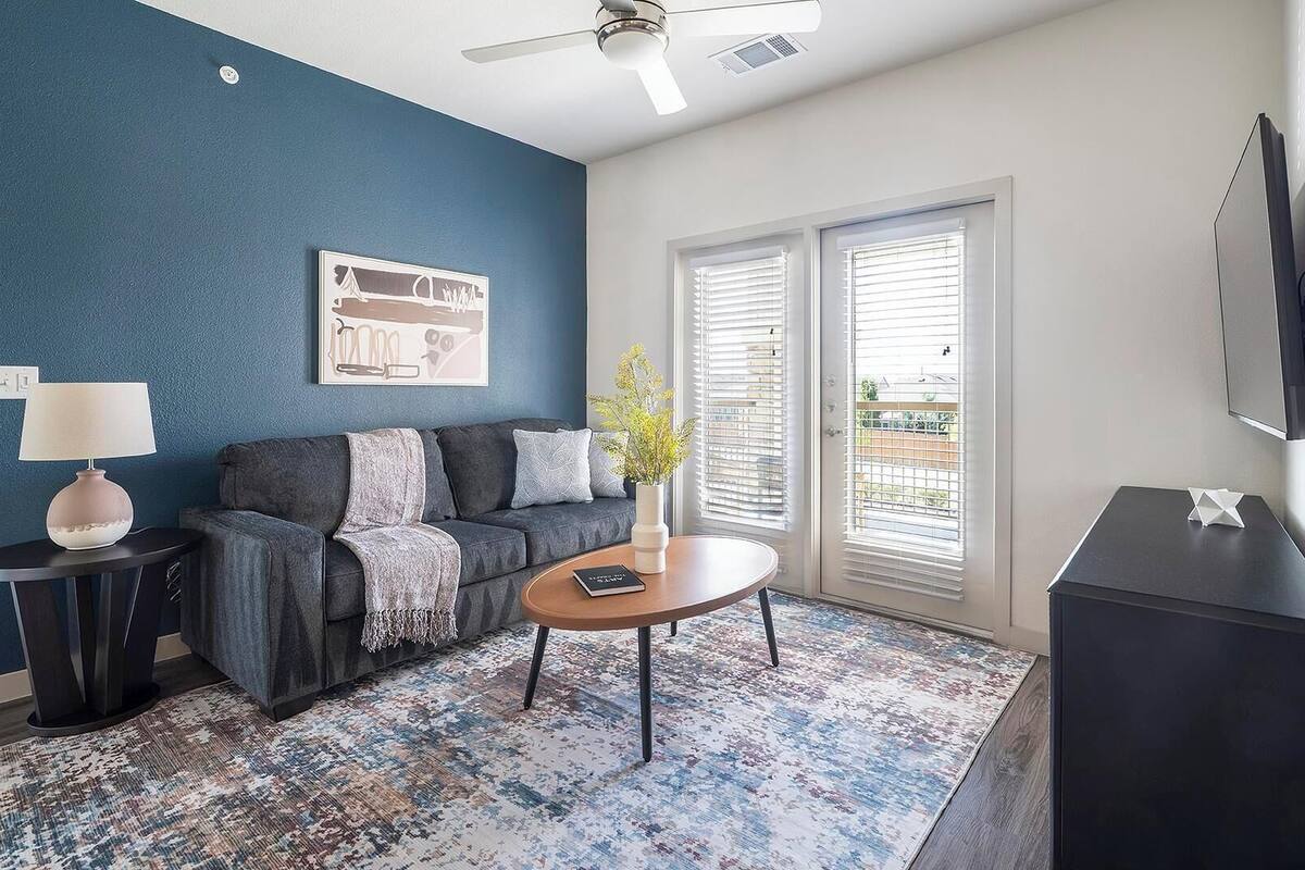 Un salon avec un mur d’accent bleu, un canapé gris, une table basse, une lampe, un tableau, un vase avec des fleurs, un téléviseur et une fenêtre avec des stores.