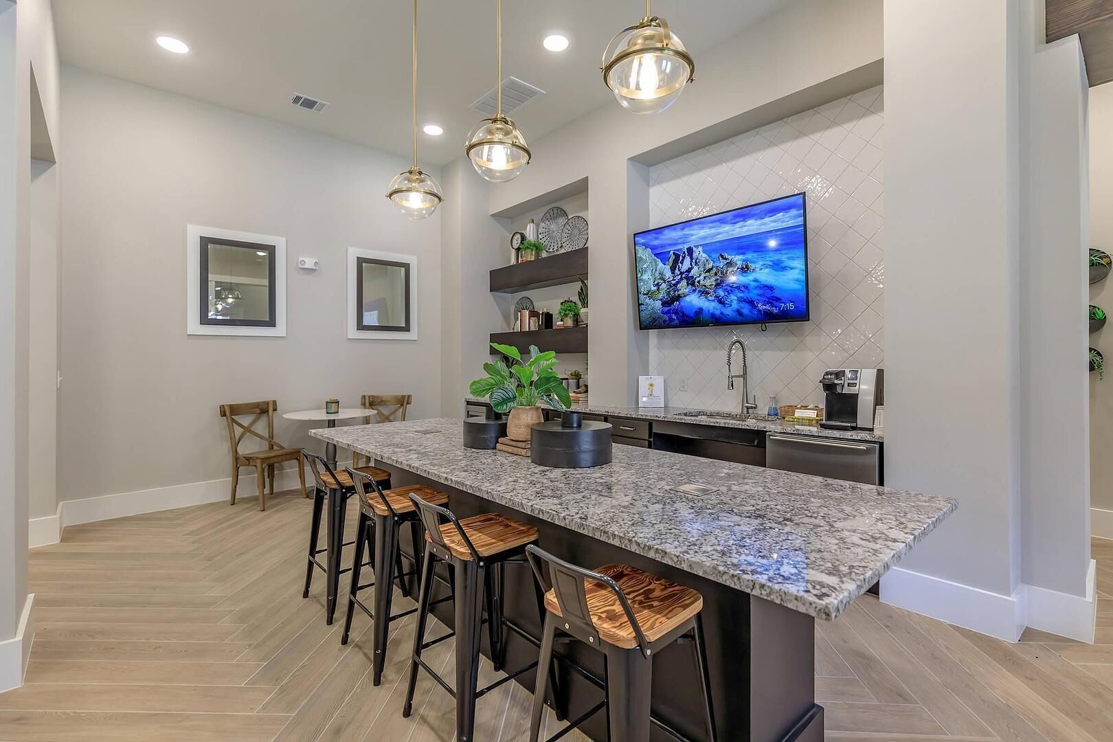 A modern kitchen with a granite island, bar stools, pendant lights, and a flat-screen TV.
