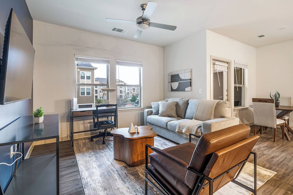 Un salon moderne avec un canapé, une table basse et un fauteuil en cuir. On y trouve également un coin repas avec une table et des chaises, ainsi qu’un ventilateur de plafond.