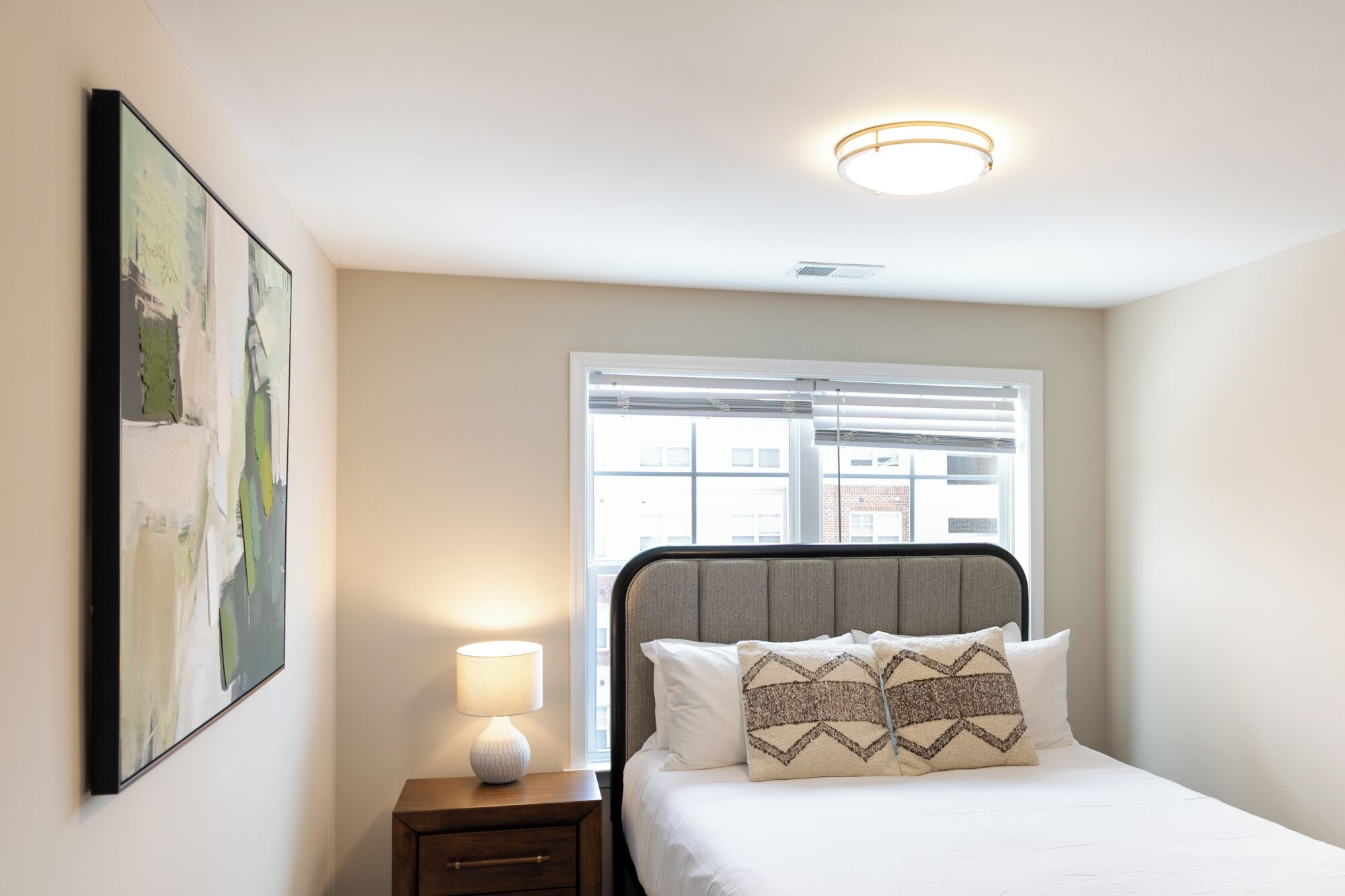 Quarto com cama, mesa de cabeceira, luminária, janela e um quadro na parede.