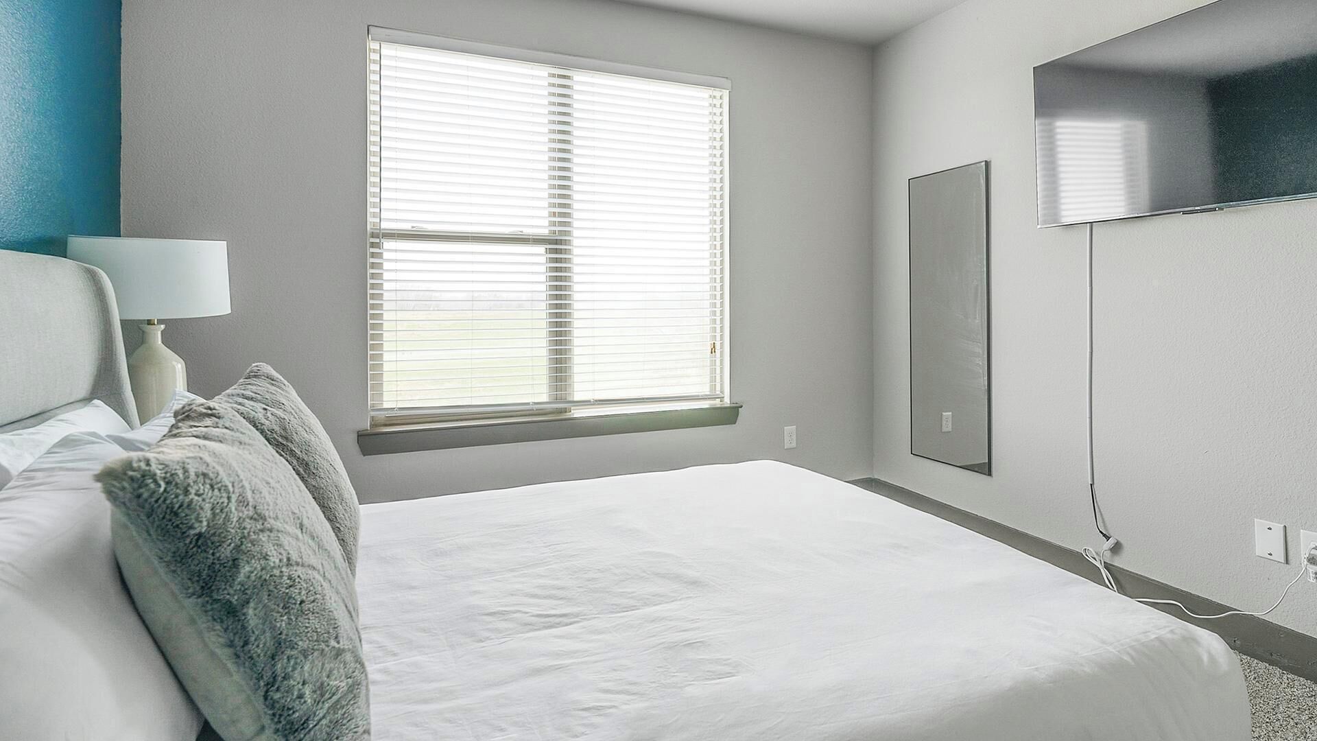 A bedroom with a bed, a window with blinds, a bedside lamp, and a wall-mounted TV.