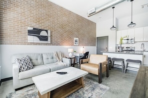 A modern living room with a sofa, coffee table, and a chair. There is a kitchen area with a stove and cabinets.