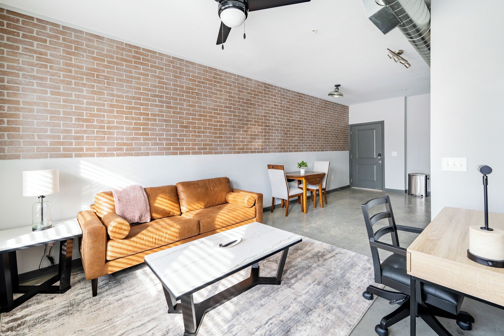 Un salon moderne avec un mur en briques, un canapé, une table basse, une table à manger avec des chaises et un ventilateur de plafond.
