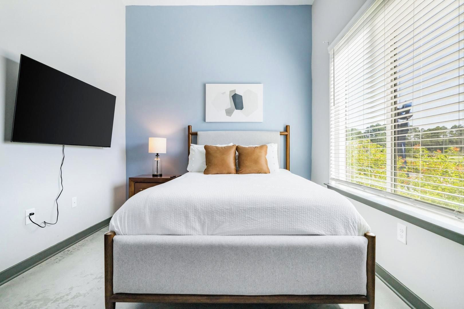 A bedroom with a bed, a bedside table with a lamp, a flat-screen TV mounted on the wall, and a window with blinds.