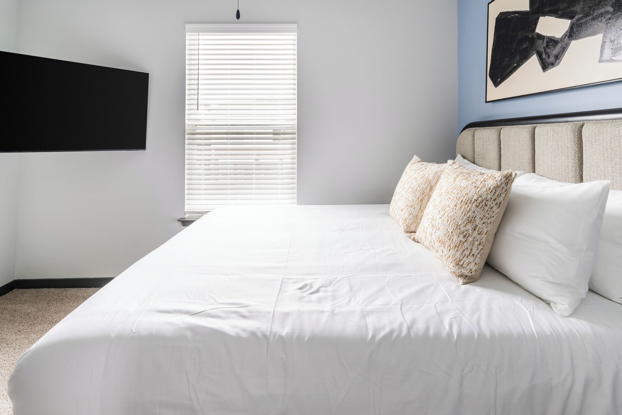 A bedroom with a large bed, white bedding, a headboard with two pillows, a window with blinds, and a wall-mounted TV.