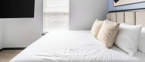 A bedroom with a large bed, white bedding, a headboard with two pillows, a window with blinds, and a wall-mounted TV.