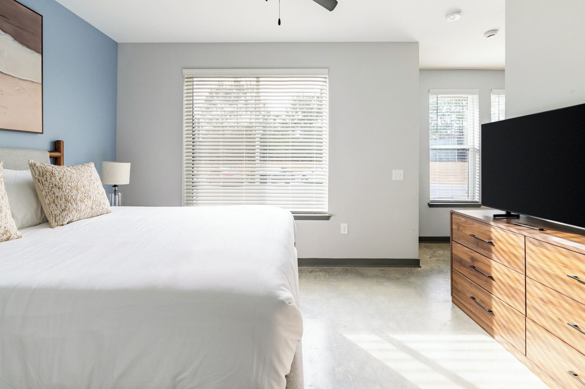 A bedroom with a bed, a TV on a wooden dresser, and a window with blinds.