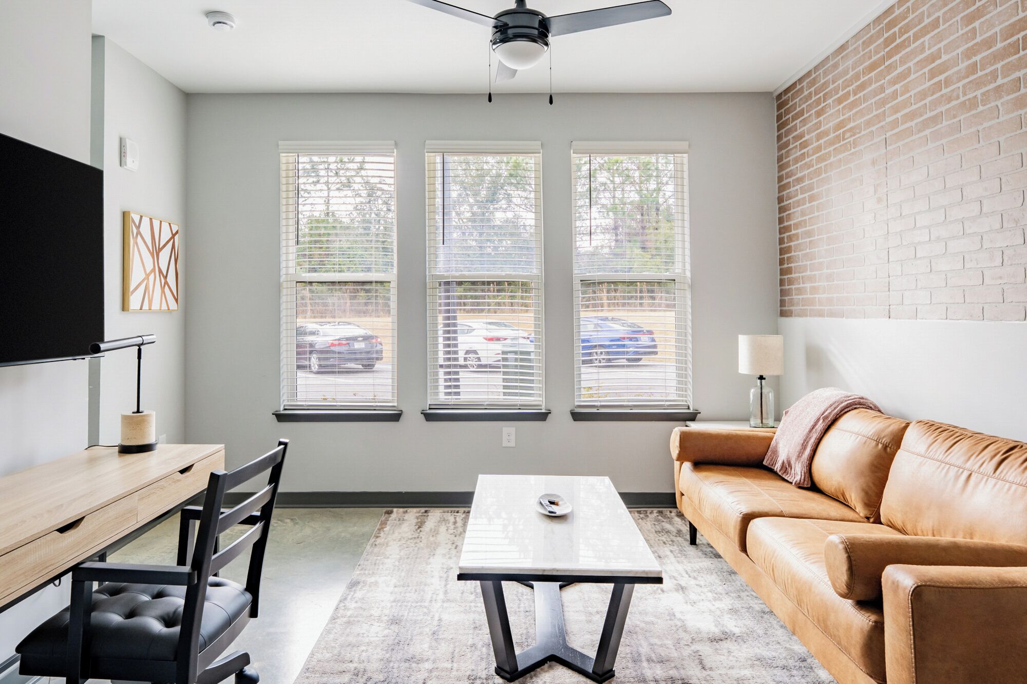 A modern living room with a wooden desk, a leather couch, and a white table. There are three windows with blinds, a ceiling fan, and a brick wall.